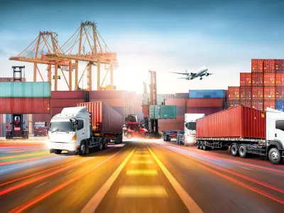 Caminhões transportando contêineres em terminal logístico de exportação, com porto, guindastes, empilhadeiras e avião ao fundo, representando transporte internacional de cargas e comércio exterior.