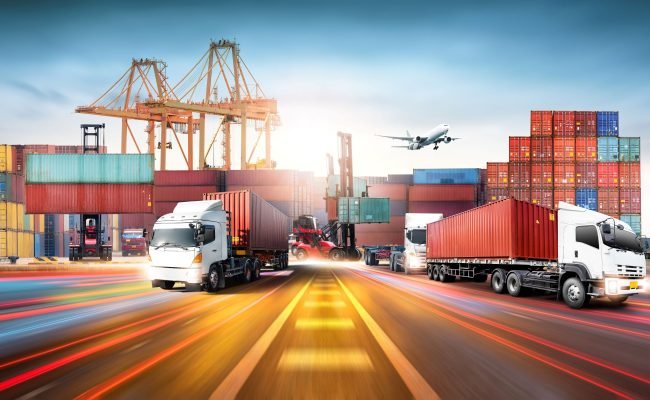 Caminhões transportando contêineres em terminal logístico de exportação, com porto, guindastes, empilhadeiras e avião ao fundo, representando transporte internacional de cargas e comércio exterior.